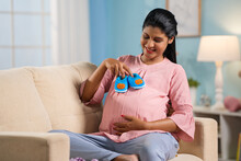 Happy Indian Pregnant Woman Playing With Baby Shoes Or Booties On Tummy At Home - Concept Of Prenatal Connection, Motherhood And Pregnancy Joy