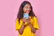 © antianti - Young cute cheerful Indian woman with phone looks away coming up SMS message for boyfriend or comment for blog in social network stands on isolated pink background. Blogger, influencer