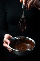 Wall Mural - A bowl of melted chocolate and a kitchen whisk are held by hands. Kitchen utensils. On a black background.