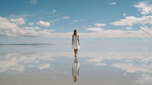 Young Woman In White Dress Standing On The Salt Lake With Reflection In Water And Looking At Blue Sky. Woman In White Dress Standing On The Salt Lake With Smooth Surface.   AI Generated