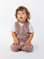 A cute curly-haired toddler boy of 1-2 years old sits on the floor with his hands folded on his knees and looks attentively with his mouth open. Copy space. Photo.