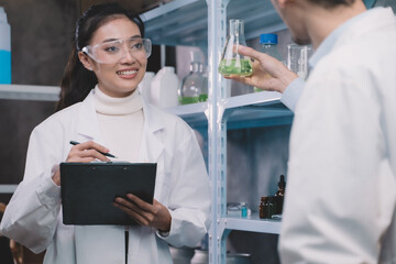 Wall Mural - Young researcher woman is note on a pen in notebook and looking a simples liquid in tube at hand of a researcher man in the laboratory.