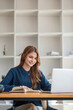 © wichayada - Smiling asian undergraduate teen girl student study in library with laptop books doing online research for coursework, making notes for essay homework assignment, online education e-learning concept