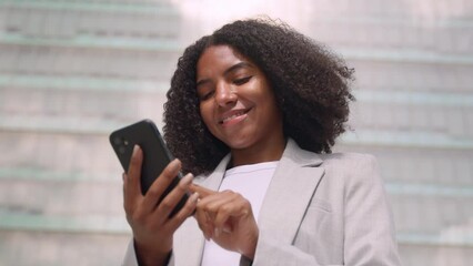 Wall Mural - Happy busy young African American business woman using mobile phone outdoors. Smiling female professional holding smartphone standing at city street building looking at cell working outside office.