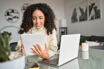 Wall Mural - Young happy latin woman, smiling curly casual girl student using digital tablet and laptop elearning or hybrid working at home online looking at tab technology device sitting at table in living room.