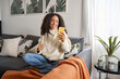 © insta_photos - Smiling latin girl sitting on couch using cell phone in living room at home. Relaxed happy young woman holding mobile cellphone technology doing shopping, communicating online, watching social media.