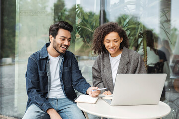 Confident smiling university students studying, learning language using laptop computer, online education concept. Successful Indian man and African American woman working together, planning startup