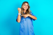 Young Caucasian Blonde Doctor Woman Wearing Blue Uniform In Hurry Pointing To Wrist Watch, Impatience, Looking At The Camera With Relaxed Expression
