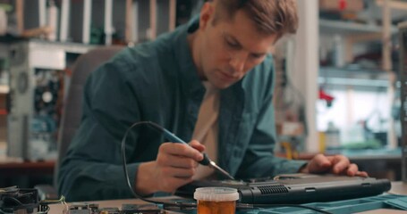 Wall Mural - young man working with broken disassembled laptop. Electronic components. Computer analysis. Slow motion attractive man using a soldering iron to fix broken hardware at the repair shop.