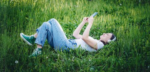 Wall Mural - Young cheerful woman in a white t-shirt lying on the green grass in the garden looking into the smartphone screen. Summer lifestyle
