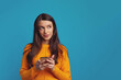 Dreamy young good looking girl holds mobile phone has thoughtful expression thinks about received message looks away isolated over blue background