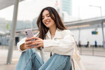 bright and cheerful young woman with shopper on shoulder waiting public transport at station smiling