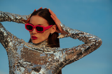 Fashionable confident woman wearing trendy fuchsia color rectangular sunglasses, sequin  turtleneck top, posing outdoor, against blue sky. Close up fashion portrait. Copy, empty space for text