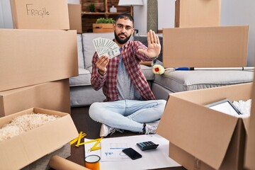 Wall Mural - Middle east man with beard sitting on the floor at new home holding money with open hand doing stop sign with serious and confident expression, defense gesture