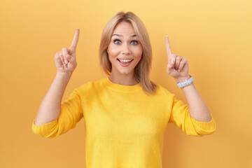 Poster - Young caucasian woman wearing yellow sweater smiling amazed and surprised and pointing up with fingers and raised arms.