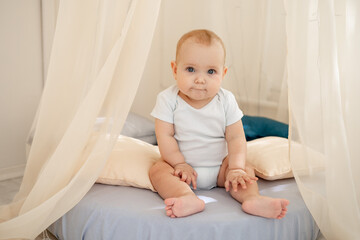 Wall Mural - cute little baby boy in a bright children's room in a wigwam or bed in a white cotton bodysuit playing