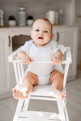 Wall Mural - a happy baby in a white cotton bodysuit on a high chair in a bright kitchen wants to eat, a small smiling baby boy or girl