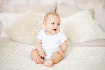 Wall Mural - a baby boy or girl of six months is sitting at home on a bed in a bright bedroom and smiling or laughing, a happy newborn in a white bodysuit