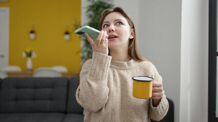 Wall Mural - Young blonde woman talking on smartphone drinking coffee at home