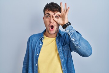 Sticker - Young hispanic man standing over blue background doing ok gesture shocked with surprised face, eye looking through fingers. unbelieving expression.