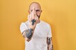 Young hispanic man with tattoos standing over yellow background showing middle finger, impolite and rude fuck off expression