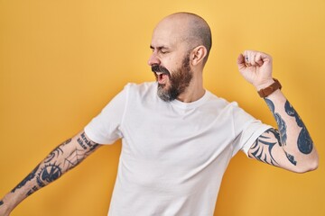 Poster - Young hispanic man with tattoos standing over yellow background dancing happy and cheerful, smiling moving casual and confident listening to music