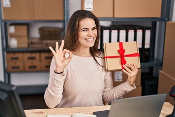 Sticker - Beautiful woman working at small business ecommerce holding gift doing ok sign with fingers, smiling friendly gesturing excellent symbol