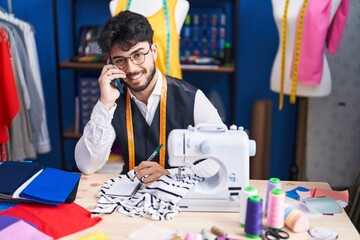 Poster - Young hispanic man tailor talking on smartphone writing on notebook at sewing studio