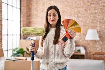 Poster - Young brunette woman holding roller painter and paint samples relaxed with serious expression on face. simple and natural looking at the camera.