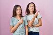 Young mother and daughter standing over pink background pointing with hand finger to the side showing advertisement, serious and calm face