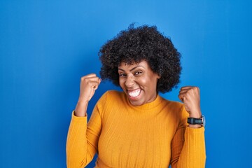 Poster - Black woman with curly hair standing over blue background celebrating surprised and amazed for success with arms raised and eyes closed. winner concept.