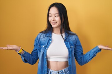 Poster - Young asian woman standing over yellow background smiling showing both hands open palms, presenting and advertising comparison and balance