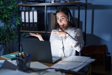 Sticker - Young brunette woman wearing call center agent headset working late at night amazed and smiling to the camera while presenting with hand and pointing with finger.