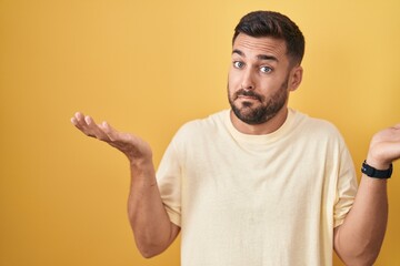 Poster - Handsome hispanic man standing over yellow background clueless and confused expression with arms and hands raised. doubt concept.