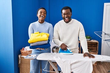 Sticker - Young african american couple ironing clothes at laundry room smiling and laughing hard out loud because funny crazy joke.