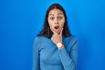 Sticker - Young brazilian woman standing over blue isolated background looking fascinated with disbelief, surprise and amazed expression with hands on chin