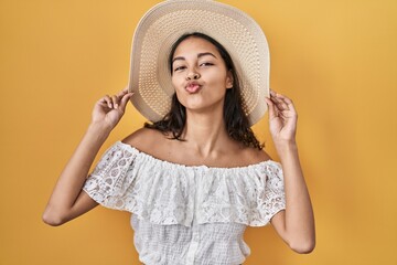 Wall Mural - Young brazilian woman wearing summer hat over yellow background looking at the camera blowing a kiss being lovely and sexy. love expression.