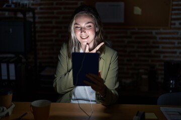 Poster - Blonde caucasian woman working at the office at night cheerful with a smile on face pointing with hand and finger up to the side with happy and natural expression