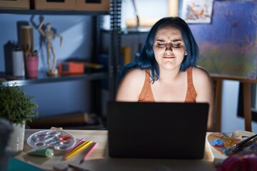 Sticker - Young modern girl with blue hair sitting at art studio with laptop at night with hand on stomach because indigestion, painful illness feeling unwell. ache concept.