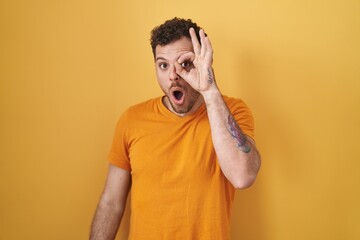 Sticker - Young hispanic man standing over yellow background doing ok gesture shocked with surprised face, eye looking through fingers. unbelieving expression.