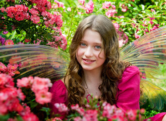 little beautiful blonde girl in pink dress with fairy wings sitting on green grass in roses garden on sunny summer day