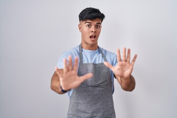 Poster - Hispanic young man wearing apron over white background afraid and terrified with fear expression stop gesture with hands, shouting in shock. panic concept.