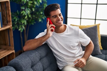 Poster - Young hispanic man talking on the smartphone sitting on sofa at home