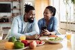 © Goran - Happy African American couple using a phone while having breakfast in the morning. Focus is on woman
