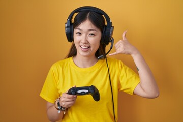 Wall Mural - Chinese young woman playing video game holding controller smiling doing phone gesture with hand and fingers like talking on the telephone. communicating concepts.