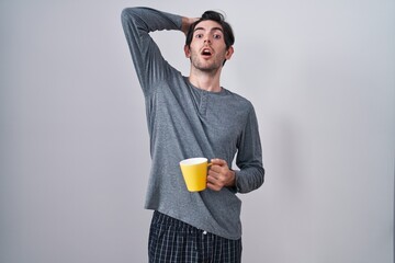 Young hispanic man wearing pajama drinking a cup of coffee crazy and scared with hands on head, afraid and surprised of shock with open mouth