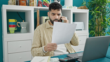 Sticker - Young hispanic man reading document talking on smartphone at dinning room