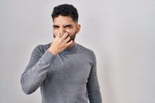 Hispanic Man With Beard Standing Over White Background Smelling Something Stinky And Disgusting, Intolerable Smell, Holding Breath With Fingers On Nose. Bad Smell