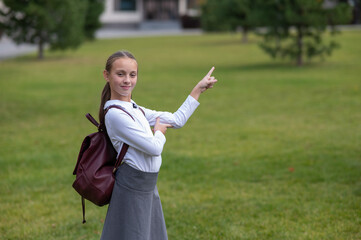 Wall Mural - Caucasian girl in uniform and with a backpack points to an empty place outdoors after school.