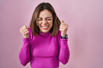 Wall Mural - Hispanic woman standing over pink background excited for success with arms raised and eyes closed celebrating victory smiling. winner concept.